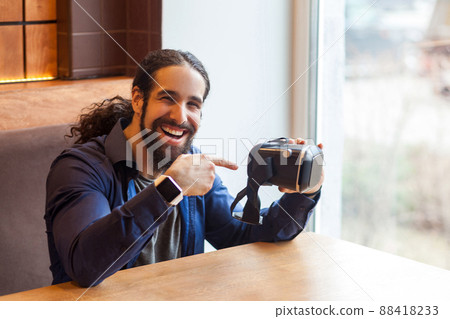 Portrait of happy bearded young adult man in casual style sitting in cafe, holding vr and pointing finger on simulator with happy face, looking at camera and toothy smile. Indoor, lifestyle concept 88418233
