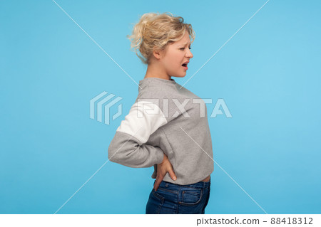 Portrait of overworked unhealthy adult woman with curly hair in sweatshirt suffering back pain, suffering lower lumbar discomfort, injured spine disk. indoor studio shot isolated on blue background 88418312