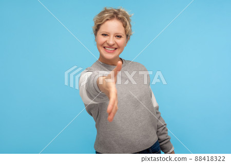 Nice to meet you. Hospitable woman with short hair in casual sweatshirt smiling friendly at camera and giving hand to handshake, trust and agreement. indoor studio shot isolated on blue background 88418322