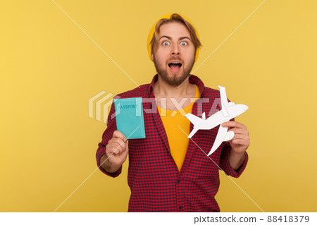 Amazed hipster tourist guy in beanie hat, checkered shirt holding paper plane and passport, shocked by tour abroad, visa free travelling by air transportation. studio shot isolated, yellow background 88418379