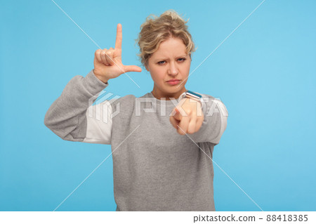 You are loser. Young woman with short curly hair in sweatshirt showing lame or loser gesture pointing to camera, blaming accusing for defeat, your fault. indoor studio shot isolated on blue background 88418385
