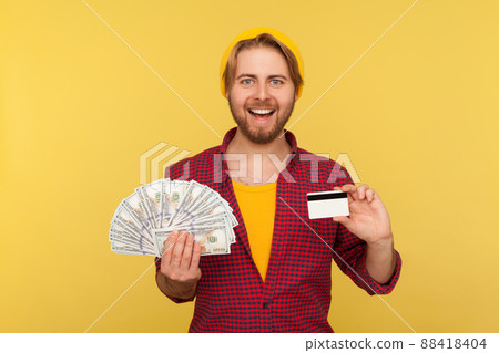 Bank loan, financial savings and commerce. Cheerful hipster guy in checkered shirt holding dollar bills and credit card, looking at camera with toothy smile. studio shot isolated on yellow background Bank loan, financial savings and commerce. Cheerful hipster guy in checkered shirt holding dollar bills and credit card, looking at camera with toothy smile. studio shot isolated on yellow background 88418404
