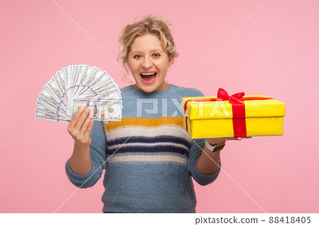 Money and gift. Portrait of shocked excited woman with curly hair in warm sweater holding present box and dollar bills, surprised by bonuses, cashback. indoor studio shot isolated on pink background 88418405
