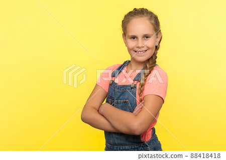 Portrait of cheerful little girl with braid in denim overalls holding hands crossed and smiling at camera, happy child expressing confidence optimism. indoor studio shot isolated on yellow background Portrait of cheerful little girl with braid in denim overalls holding hands crossed and smiling at camera, happy child expressing confidence optimism. indoor studio shot isolated on yellow background 88418418