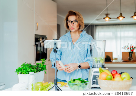 Middle age women is blogging for her wellness lifestyle channel about healthy balanced living in the kitchen of her home. Creating video content for social media with phone camera on a tripod. 88418697