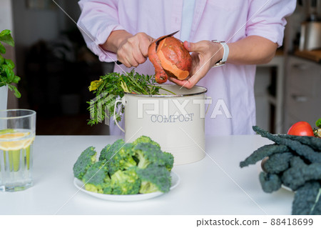 Compost the kitchen waste, recycling at home. Woman cleaning sweet potato and putting vegetables cutted leftovers into the garbage, compost bin on her kitchen. Environmentally responsible, ecology 88418699