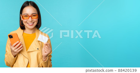 Online shopping. Stylish young asian woman in sunglasses, showing credit card and using smartphone, paying in internet, making purchase, standing over blue background 88420139