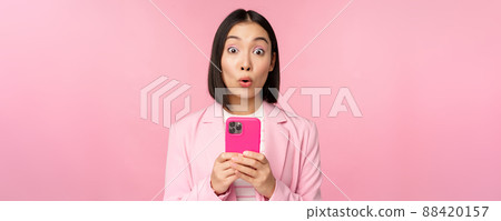 Portrait of asian businesswoman with surprised face, using smartphone app, wearing business suit. Korean girl with mobile phone and excited face expression, pink background 88420157