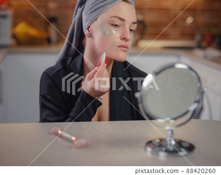young woman looks in mirror using facial gua sha jade board at home. young woman looks in mirror using facial gua sha jade board at home. 88420260