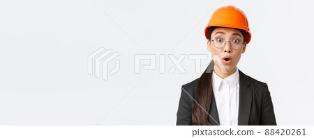 Close-up of shocked and concerned asian female engineer hear bad news, worrying announcement, standing in safety mask, glasses and business suit and gasping startled, white background 88420261