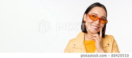 Close up portrait of asian woman thinking, wearing sunglasses and smiling, looking up thoughtful, standing over white studio background 88420510