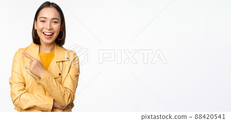 Portrait of beautiful korean girl laughing, smiling and pointing finger left at logo, showing banner, standing in casual outfit over white background 88420541