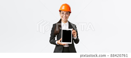Smiling professional female engineer, saleswoman or estate agent showing location to client, standing in safety helmet and business suit, introduce construction project at tablet screen Smiling professional female engineer, saleswoman or estate agent showing location to client, standing in safety helmet and business suit, introduce construction project at tablet screen 88420761