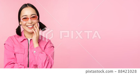 Close up portrait of stylish asian woman in sunglasses, smiling, looking flattered, laughing coquettish, standing over pink background 88421028