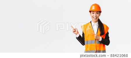 Building, construction and industrial concept. Smiling female engineer in helmet and reflective clothing showing way, pointing upper left corner, talking about constructing project, white background Building, construction and industrial concept. Smiling female engineer in helmet and reflective clothing showing way, pointing upper left corner, talking about constructing project, white background 88421068