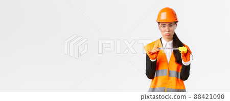 Disappointed gloomy female asian construction engineer, architect showing small size on tape measure, pouting upset with measurements, standing displeased in safety helmet, white background 88421090