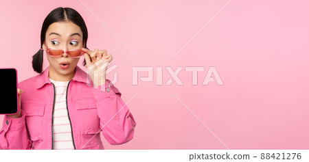 Stylish korean girl, young woman in sunglasses showing smartphone screen, mobile phone app interface or website, standing over pink background Stylish korean girl, young woman in sunglasses showing smartphone screen, mobile phone app interface or website, standing over pink background 88421276