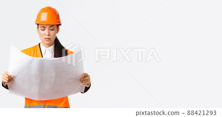 Confused and doubtful asian female chief engineer, lead architect at construction area looking displeased and hesitant at strange blueprints, reading confusing building project, white background 88421293