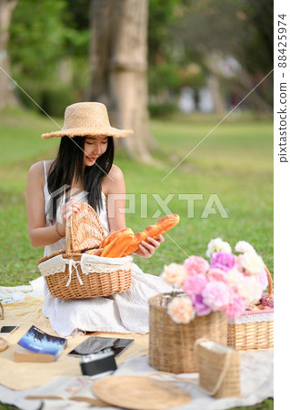 Woman with a pretty dress, wicker picnic basket with baguette and accessories on picnic cloth. 88425974