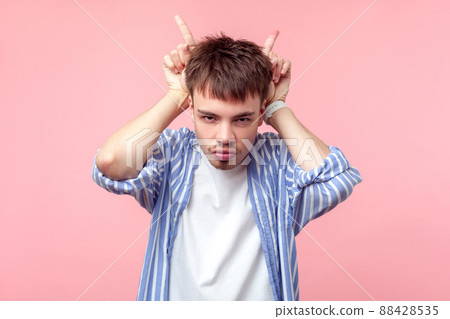 Portrait of bully young brown-haired man with small beard and mustache in casual striped shirt showing bull horn gesture with fingers, attack or defense. indoor studio shot isolated on pink background Portrait of bully young brown-haired man with small beard and mustache in casual striped shirt showing bull horn gesture with fingers, attack or defense. indoor studio shot isolated on pink background 88428535