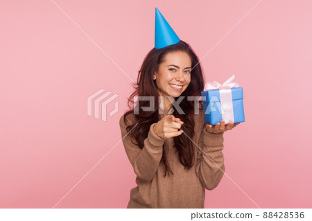 This present for you. Portrait of happy joyful young woman wearing party cone on head holding box and pointing to camera, holiday tradition of gift giving. studio shot isolated on pink background 88428536