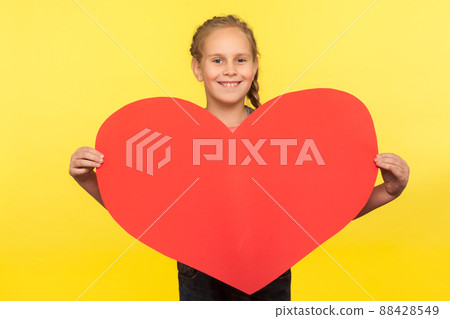 Mother's day greetings. Portrait of cheerful little girl holding big red heart and smiling to camera, child expressing love and fondness for parents. indoor studio shot isolated on yellow background Mother's day greetings. Portrait of cheerful little girl holding big red heart and smiling to camera, child expressing love and fondness for parents. indoor studio shot isolated on yellow background 88428549