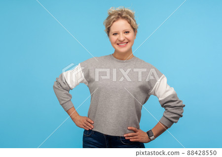Happy woman with short curly hair in sweatshirt smiling at camera and holding hands on hips, expressing optimism confidence and joy, positive emotions. indoor studio shot isolated on blue background Happy woman with short curly hair in sweatshirt smiling at camera and holding hands on hips, expressing optimism confidence and joy, positive emotions. indoor studio shot isolated on blue background 88428550
