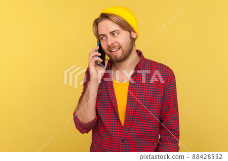 Cheerful trendy guy in hipster beanie hat and checkered shirt calling, smiling while talking mobile phone, having positive conversation on telephone. indoor studio shot isolated on yellow background Cheerful trendy guy in hipster beanie hat and checkered shirt calling, smiling while talking mobile phone, having positive conversation on telephone. indoor studio shot isolated on yellow background 88428552