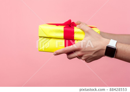 Closeup of young male hands with wristwatches giving yellow gift box with red ribbon, sharing present on holiday, concept of charity and generosity. indoor studio shot isolated on pink background Closeup of young male hands with wristwatches giving yellow gift box with red ribbon, sharing present on holiday, concept of charity and generosity. indoor studio shot isolated on pink background 88428553