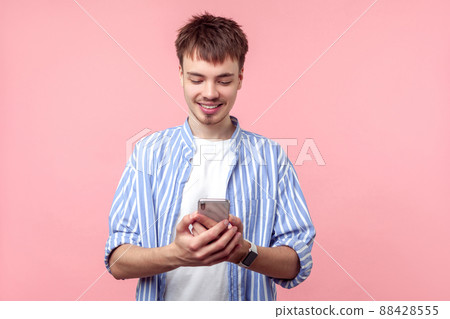 Portrait of positive brown-haired man with small beard and mustache in casual striped shirt holding cellphone, smiling while reading message, good news. indoor studio shot isolated on pink background Portrait of positive brown-haired man with small beard and mustache in casual striped shirt holding cellphone, smiling while reading message, good news. indoor studio shot isolated on pink background 88428555