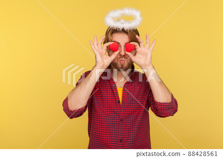 Romantic bearded guy saint halo covering eyes with toy hearts as if looking with love, sending air kiss, concept of affection adoration in relations. indoor studio shot isolated on yellow background 88428561
