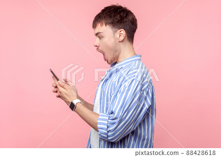 Side view of amazed brunette man with small beard and mustache in casual striped shirt holding phone, reading message with open mouth, astonished face. indoor studio shot isolated on pink background Side view of amazed brunette man with small beard and mustache in casual striped shirt holding phone, reading message with open mouth, astonished face. indoor studio shot isolated on pink background 88428618