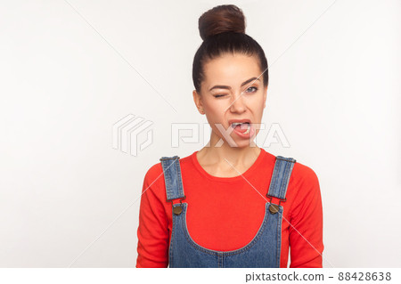 Portrait of funny displeased girl with hair bun in denim overalls winking to camera, looking with ugly skewed face, nasty dissatisfied grimace, expressing displeasure. indoor studio shot, isolated 88428638