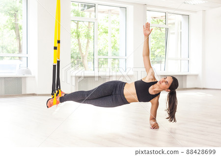 Portrait of young attractive muscular woman in black top and leggings standing on perfect side plank position use fitness straps, doing push ups while legs hanging on trx. Indoor, window background 88428699