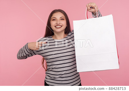 Portrait of joyful lovely woman in striped sweatshirt pointing shopping bag and looking at camera with toothy smile, showing package with blank space for advertise. indoor studio shot, pink background Portrait of joyful lovely woman in striped sweatshirt pointing shopping bag and looking at camera with toothy smile, showing package with blank space for advertise. indoor studio shot, pink background 88428713