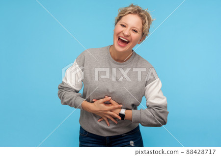 Happiness and laughter. Extremely excited woman with short hair in sweatshirt holding belly and laughing out loudly, cracking up, amused by funny joke. indoor studio shot isolated on blue background 88428717