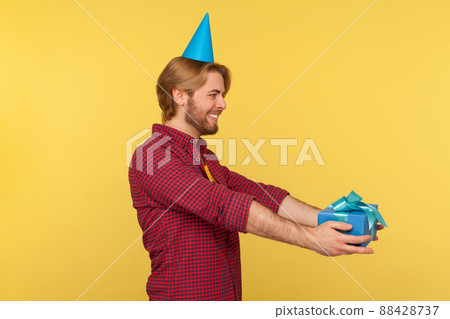Side view, cheerful delighted guy in funny party cone holding wrapped present, giving gift box greeting happy birthday, congratulating on anniversary. studio shot isolated on yellow background 88428737