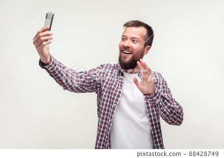 Hello. Portrait of positive handsome bearded man in casual plaid shirt talking on video call using smartphone and waving hand making hi gesture, mobile communication. studio shot, white background 88428749