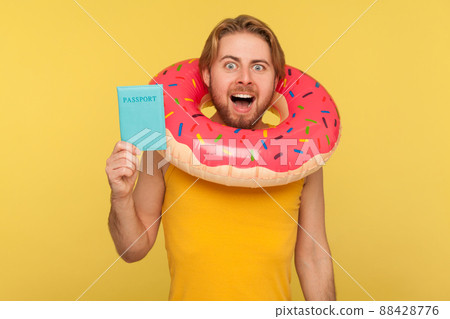 Happy optimistic funny tourist guy in undershirt standing with pink donut rubber ring around head and holding passport document, visa free travel abroad. studio shot isolated on yellow background Happy optimistic funny tourist guy in undershirt standing with pink donut rubber ring around head and holding passport document, visa free travel abroad. studio shot isolated on yellow background 88428776
