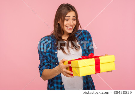 Surprising bonus, holiday present. Portrait of amazed happy girl in checkered shirt holding wrapped box and smiling, looking excited and shocked by sudden gift. studio shot isolated on pink background 88428839