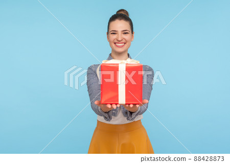 Attractive joyful fashionably dressed woman with hair bun giving gift box and smiling broadly, holding out present to camera, congratulating on birthday. indoor studio shot isolated on blue background 88428873