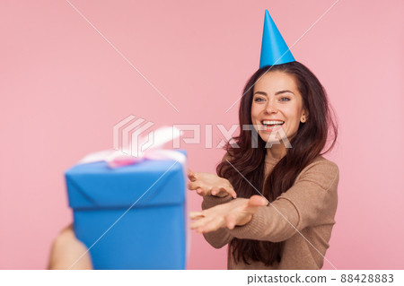 Portrait of happy cheerful young woman with party cone on head outstretching hands to take gift box and laughing, expressing great joy about birthday present. studio shot isolated on pink background 88428883