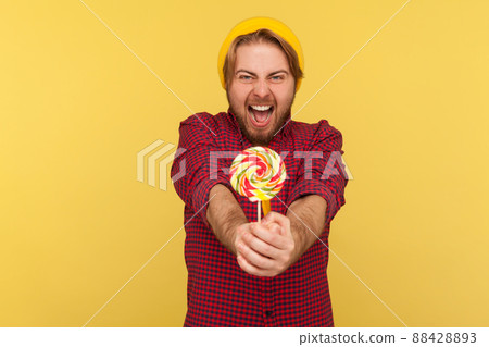 Extremely joyful delighted hipster guy in beanie hat, checkered shirt giving lollipop to camera and screaming with excitement, crazy about sweet sugary confectionery. studio shot isolated on yellow Extremely joyful delighted hipster guy in beanie hat, checkered shirt giving lollipop to camera and screaming with excitement, crazy about sweet sugary confectionery. studio shot isolated on yellow 88428893
