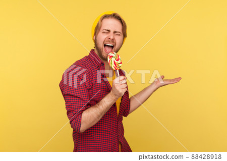 Amazing hipster guy in beanie hat and checkered shirt screaming into lollipop like microphone, pretending to sing loudly in karaoke , having fun. indoor studio shot isolated on yellow background Amazing hipster guy in beanie hat and checkered shirt screaming into lollipop like microphone, pretending to sing loudly in karaoke , having fun. indoor studio shot isolated on yellow background 88428918