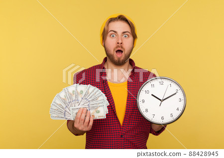 Wow, time to make money. Amazed hipster bearded guy in checkered shirt holding big clock and dollars banknotes, looking at camera shocked surprised. indoor studio shot isolated on yellow background Wow, time to make money. Amazed hipster bearded guy in checkered shirt holding big clock and dollars banknotes, looking at camera shocked surprised. indoor studio shot isolated on yellow background 88428945