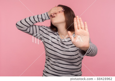 No, don't want to look at this. Portrait of woman in striped sweatshirt covering eyes and turning with disgust from something shameful, showing stop gesture. studio shot isolated on pink background No, don't want to look at this. Portrait of woman in striped sweatshirt covering eyes and turning with disgust from something shameful, showing stop gesture. studio shot isolated on pink background 88428962