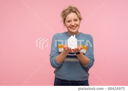 Portrait of positive adult woman with curly hair in sweater holding paper house on palms and smiling at camera, home purchase, property insurance and investment. indoor studio shot pink background Portrait of positive adult woman with curly hair in sweater holding paper house on palms and smiling at camera, home purchase, property insurance and investment. indoor studio shot pink background 88428975