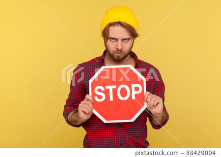 Determined serious hipster guy in beanie hat and checkered shirt holding traffic Stop sign and looking menace, warning of road safety, pedestrian crossing. studio shot isolated on yellow background 88429004