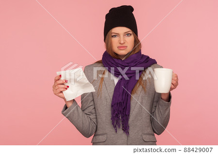 Portrait of irritated flu-sick woman in warm hat and scarf holding tissue and cup of hot beverage, looking with annoyed expression, treating influenza. indoor studio shot isolated on pink background Portrait of irritated flu-sick woman in warm hat and scarf holding tissue and cup of hot beverage, looking with annoyed expression, treating influenza. indoor studio shot isolated on pink background 88429007
