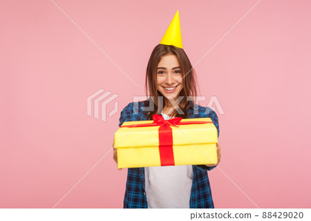 Take it, this present for you. Portrait of happy brunette girl with party cone on head giving gift box to camera and smiling, congratulating on holiday. indoor studio shot isolated on pink background 88429020